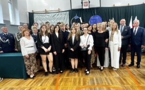 Grupa abiturientów ZS CKZ Bujny wraz z Dyrekcją, wychowawcami i zaproszonymi Gośćmi. Sala gimnastyczna, w tle na zielonym suknie hasło z białych liter oraz granatowe dekoracje związane z edukacją. Częściowo widoczne też stojaki z flagami Polski. 