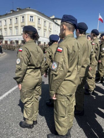 Uczniowie klas mundurowych ZS CKZ Bujny w umundurowaniu i nakryciach głowy podczas obchodów rocznicy uchwalenia Konstytucji 3 Maja. W tle inni uczestnicy uroczystości oraz zabudowania.