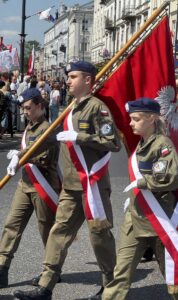 Poczet sztandarowy ZS CKZ Bujny. Uczniowie klas mundurowych OPW w umundurowaniu z logo szkoły oraz flagą polski, nakryciach głowy, białych rękawiczkach i w szarfach w barwach narodowych idą ze sztandarem. W tle zabudowania oraz pozostali uczestnicy obchodów rocznicy uchwalenia Konstytucji 3. Maja. 