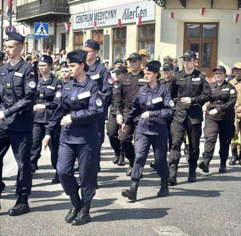 Uczniowie klas mundurowych idą w defiladzie podczas uroczystości odchodów rocznicy uchwalenia Konstytucji 3 Maja. W tle zabudowania przystrojone flagami Polski.