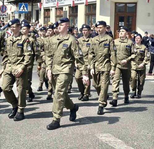 Uczniowie klas mundurowych idą w defiladzie podczas uroczystości odchodów rocznicy uchwalenia Konstytucji 3 Maja. W tle zabudowania przystrojone flagami Polski.