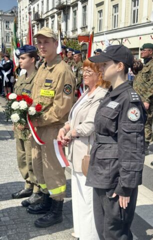 Delegacja szkoły, 3 uczniów klas mundurowych w umundurowaniu oraz nakryciach głowy wraz z p. Wicedyrektor. Stoją do złożenia kwiatów. Uczeń trzyma w ręku bukiet z białych i czerwonych goździków z szarfą w barwach narodowych, p. wicedyrektor trzyma w ręku małą flagę Polski.