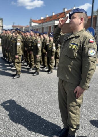 Uczeń klasy mundurowej w umundurowaniu oraz nakryciu głowy na obchodach rocznicy uchwalenia Konstytucji 3 Maja. Salutuje. W tle ustawieni w szeregach uczestnicy obchodów.