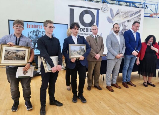 Laureaci i finaliści konkursu historycznego oraz organizatorzy wewnątrz sali gimnastycznej. W tle baner reklamowy szkoły oraz po lewej stronie duży plakat konkursu. Stojąca po prawej stornie kobieta trzyma mikrofon w lewej ręce i przemawia. Zwyciezcy konkursu stoją i trzymają w ręku dyplomy, nagrody książkowe oraz pamiątkowe obrazy w ramkach.