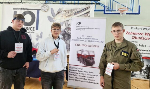 3 finalistów z Zespołu Szkół CKZ Bujny, jeden z finalistów, stojący po prawej stronie, uczeń klasy OPW w mundurze wojskowym, oliwkowym. Zwycięzcy stoją w sali gimnastycznej, na szyjach mają identyfikatory z czerwonym napisem finalista i tekstami . W tle baner reklamowy konkursu z napisem: RP Patronat Honorowy Prezydenta Rzeczpospolitej Polskiej Andrzeja Dudy, XI edycja konkursu wojewódzkiego Żołnierze Wyklęci Obudźcie Polskę Finał Wojewódzki oraz nazwiskami organizatorów.