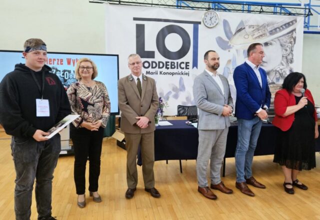 Jeden z finalistów konkursu wraz z organizatorami w sali gimnastycznej podczas ceremonii wręczania nagród. Na szyi ma zawieszony identyfikator. Po prawej stronie stoi kobieta i trzyma w prawej ręce mikrofon. W tle banery reklamowe szkoły LO Poddębice oraz plakat konkursu.