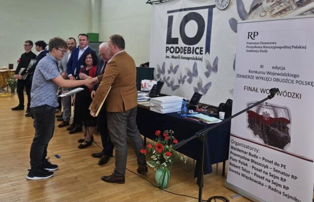Laureat konkursu odbiera nagrodę i gratulacje z rąk organizatorów konkursu w sali gimnastycznej. W tle inni finaliści konkursu. widoczne także baner szkoły LO Poddębice oraz baner konkursu i stoliki przykryte granatowym suknem, na których znajdują się nagrody dla finalistów. Przed stolikami na podłodze stoi zielony wazon z białymi i czerwonymi kwiatami.