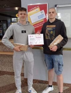Dwóch uczniów w budynku poczty. Trzymają w ręku pudełka tekturowe oraz kartkę informacyjną o akcji Góra Grosza. 