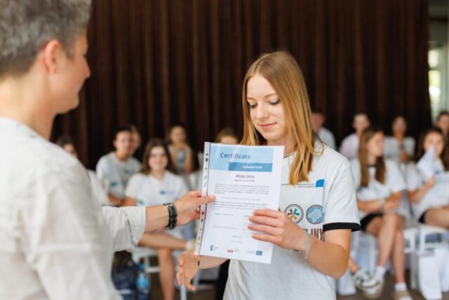 Uczestniczka zagranicznej praktyki zawodowej odbiera certyfikat i gratulacje od kobiety. W tle siedzą pozostali uczestnicy praktyk.