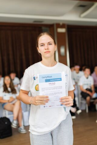 Uczestniczka zagranicznych praktyk zawodowych pozuje do zdjęcia w pomieszczeniu. Ma na sobie białą koszulkę wyjazdową, w rękach trzyma certyfikat potwierdzający odbycie praktyki. W tle siedzą pozostali uczestnicy praktyki.