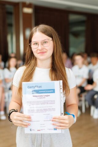 Uczestniczka zagranicznych praktyk zawodowych stoi w pomieszczeniu. w rękach trzyma certyfikat potwierdzający odbycie praktyki. W tle siedzą pozostali uczestnicy praktyk.