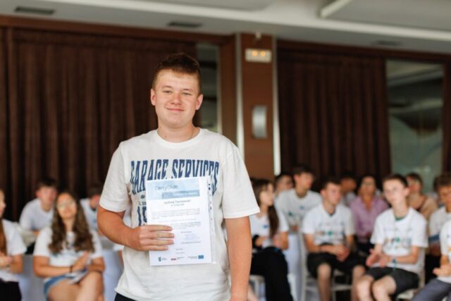 Uczestnik zagranicznych praktyk zawodowych stoi w pomieszczeniu. w rękach trzyma certyfikat potwierdzający odbycie praktyki. W tle siedzą pozostali uczestnicy praktyk.