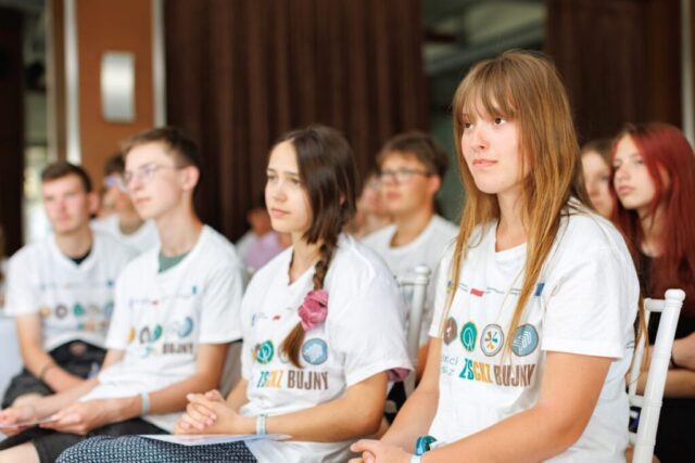 Uczestnicy zagranicznej praktyki zawodowej w białych koszulkach wyjazdowych z logo i napisem mobilności europejskie z ZS CKZ Bujny. Siedzą na białych krzesłach w pomieszczeniu.
