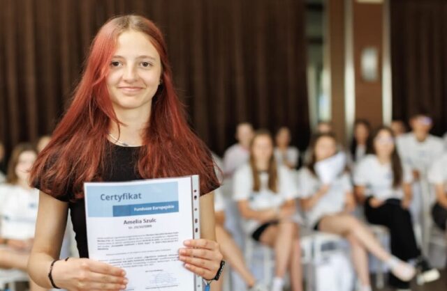 Uczestniczka zagranicznych praktyk zawodowych stoi w pomieszczeniu. W rękach trzyma certyfikat potwierdzający odbycie praktyk. W tle siedzą pozostali uczestnicy praktyk. W tle brązowa kotara.