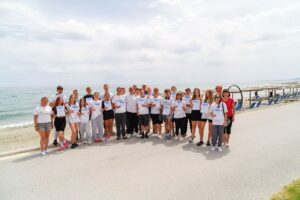 Grupa 26 uczestników zagranicznej praktyki zawodowej wraz z opiekunami na promenadzie przy plaży. Każdy z uczestników trzyma w ręku certyfikat uzyskany na zakończenie praktyki. W tle widoczne morze, słomiane parasole i granatowe leżaki, a także kilka osób. 