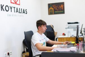 Uczestnik zagranicznych praktyk zawodowych w białej koszulce wyjazdowej siedzi w pomieszczeniu biurowym przy biurku z komputerem i pracuje nad czymś. Na ścianie nazwa i logo firmy KOYTALIAS. 