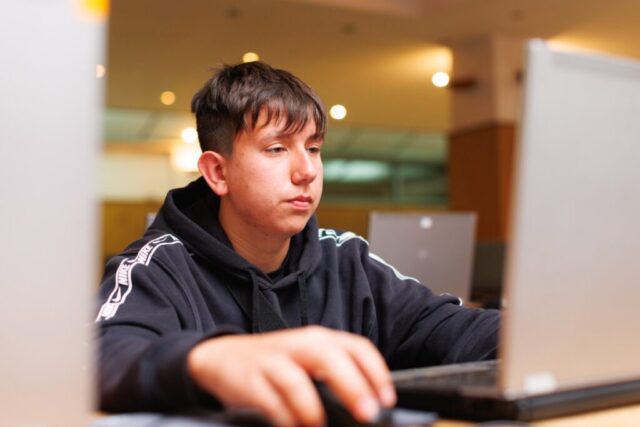Uczestnik zagranicznej praktyki zawodowej siedzi w pomieszczeniu. Wykonuje pracę na laptopie. Skupiony patrzy w ekran.