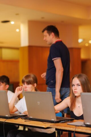Uczestnicy zagranicznej praktyki zawodowej w pomieszczeniu. Siedzą przy ławkach, na których stoją laptopy. W tle instruktor.