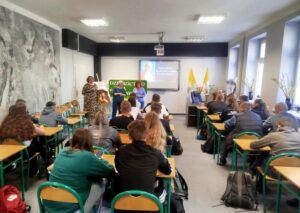 Grupa uczniów, uczestników konkursu siedzi w ławkach w dużej sali. W tle ekran, wyświetlony slajd z wizerunkiem Jana Pawła II oraz tekstem 02.04.2025r 20 rocznica śmierci św. Jana Pawła II. Klasa udekorowana wazonami z forsycją, flagami papieskimi w kolorze żółtym i białym, na sztaludze portret Papież, a także na zielonym tle napis dziedzictwo Jana Pawła II. Przed uczniami stoją organizatorki konkursu oraz p. Wicedyrektor szkoły. 