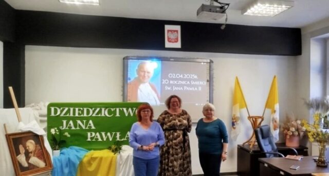 Organizatorki konkursu o Janie Pawle II w dużej klasie. W tle ekran z wyświetlonym slajdem pokazującym portret Jana Pawła II oraz napis 02.04.2025. 20 rocznica śmierci św. Jana Pawła II, widoczna także sztaluga, na której stoi portret Papieża oraz zielona tablica z napisem w białym kolorze Dziedzictwo Jana Pawła II. Klasa udekorowana kwiatami, flagami w kolorach żółtym i białym. Przed tablicą stół udekorowany materiałami w kolorach żółtym, białym i niebieskim.