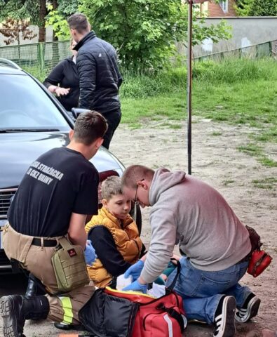 Dwóch uczestników konkursu udziela pomocy chłopcu. Uczniowie noszą niebieskie rękawice ochronne. Wszyscy są na zewnątrz. Widoczna także torba ratownicza. W tle fragment aura, dwie dorosłe osoby i fragment zabudowań oraz zieleń miejska.