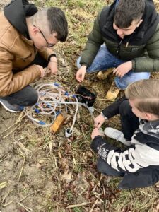 3 uczniów kuca i pobiera próbki wody. Widać aparaturę oraz wężyki laboratoryjne. 