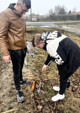 Dwóch uczniów na zewnątrz. Jeden pobiera próbki wody, drugi stoi i trzyma w ręku zestaw wężyków laboratoryjnych.
