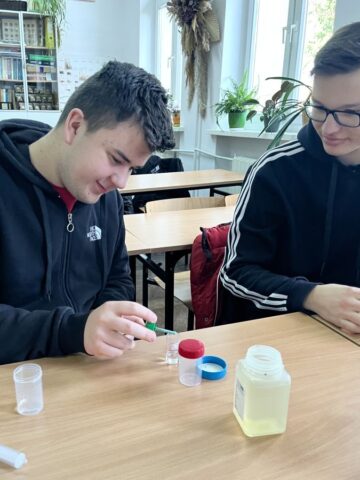 Dwóch uczniów w klasie. Siedzą w ławce i badają próbki wody. Na ławce naczynia i pojemniki laboratoryjne.