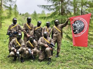 Grupa 8 uczestników obozu w strojach moro, kaskach ochronnych oraz z bronią stoi na zewnątrz na polanie w lesie. Jeden z uczestników trzyma w ręku flagę. Na czerwonym tle zarys żołnierza umundurowanego w strój maskujący i biały napis bądź niewidzialny dla wroga.