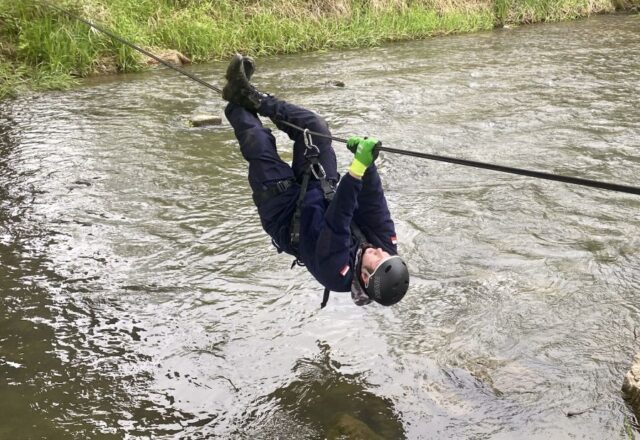 Uczestnik obozu szkoleniowego przeprawia się na linie przez rzekę. Jest przyczepiony do liny uprzężą, na głowie ma kask ochronny.