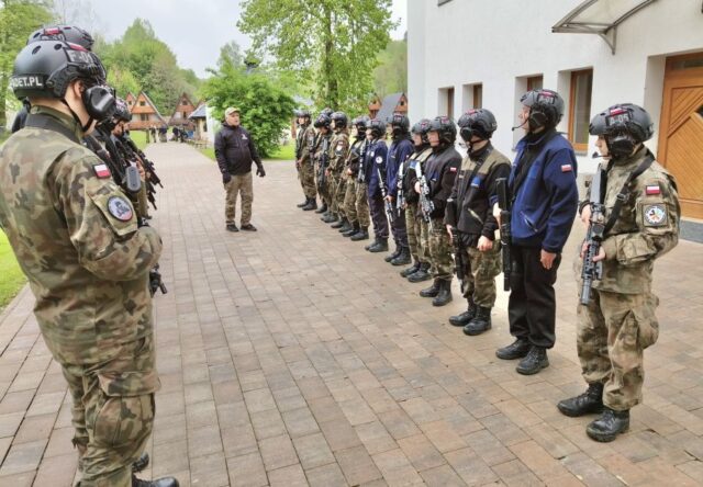 Uczestnicy obozu szkoleniowego w umundurowaniu, w kaskach z interkomem oraz z bronią podczas odprawy z instruktorami. Stoją przed budynkiem. W tle domki typu Brda i ośrodek.