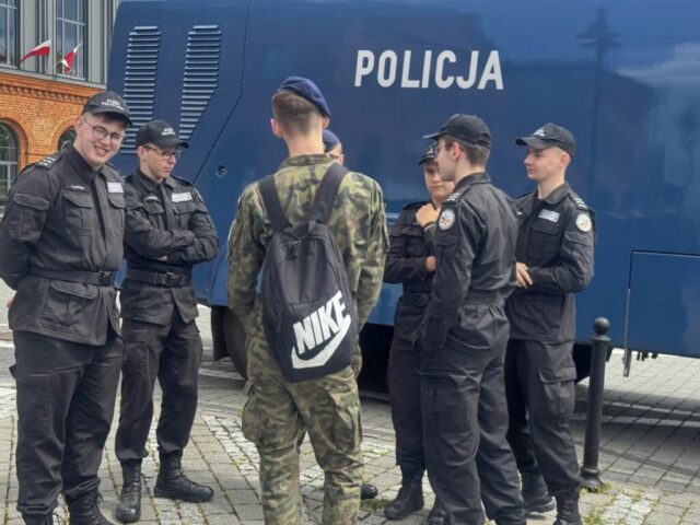 Grupa uczniów klasy policyjnej oraz opw w umundurowaniu na zewnątrz. Stoją przy granatowym wozie z napisem Policja. Po lewej stronie w tle fragment budynku z cegły.
