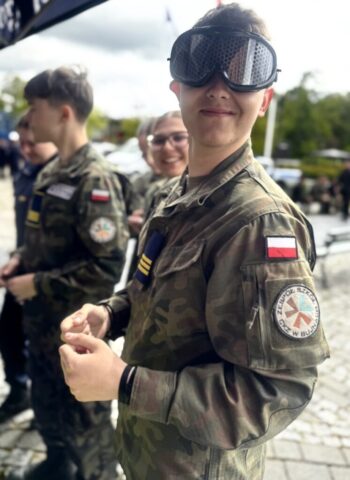 Uczeń klasy opw w umundurowaniu stoi przy stoisku. Ma na sobie okulary symulacyjne. W tle inni uczniowie w umundurowaniu.