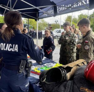 Stoisko firmowe policji na zewnątrz. Przy stoisku uczniowie w umundurowaniu. W tle grupa uczniów klasy policyjnej w umundurowaniu oraz dwóch policjantów. Widoczne także auta. 