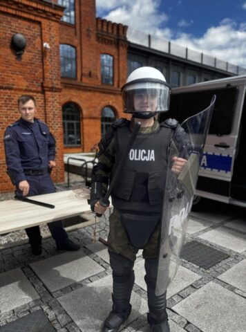 Uczeń klasy opw w umundurowaniu i ubraniu ochronnym. W ręku trzyma pałkę policyjną w a drugiej ręce ratczę. Ma na głowie kask ochronny. Po lewej stronie stoi funkcjonariusz policji w mundurze. Przed nim jest ławka na której leży pałka policyjna. W tle wóz policyjny.