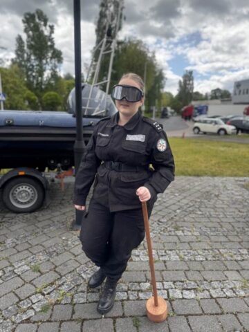 Uczennica kalsy policyjnej w umundurowaniu i okularach symulacyjnych przy pachołki. W tle zaparkowana na lawecie motorówka policyjna, fragment budynki i parking.