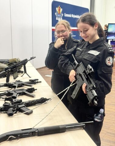 Dwie uczennice klasy policyjnej w umundurowaniu stoją przy stolikach, na których jest prezentowana broń. W tle banner reklamowy Komendy Wojewódzkiej Policji oraz fragment ekranu.