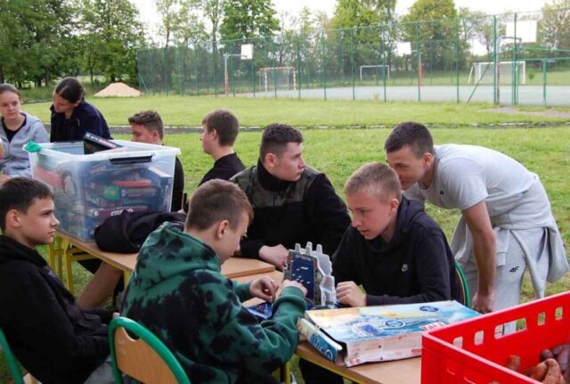 Uczniowie klas mundurowych podczas pikniku integracyjnego na terenie boiska szkolnego. Siedzą i grają w gry planszowe. W tle widoczna bieżnia oraz boisko orlik.