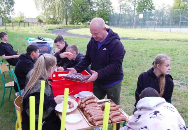 Młodzież klas mundorowych wraz z opiekunem na terenie boiska szkolnego. Młodzież siedzi przy ławkach, rozmawiają, wychowawca trzyma na tacy kiełbaski przeznaczone na ognisko. W tle boisko orlik.