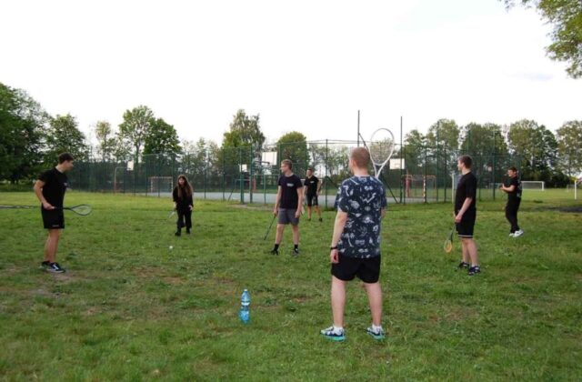 Grupa uczniów klas mundurowych na terenie boiska szkolnego. Stoją na trawie i grają w badmintona. W tle boisko orlik.