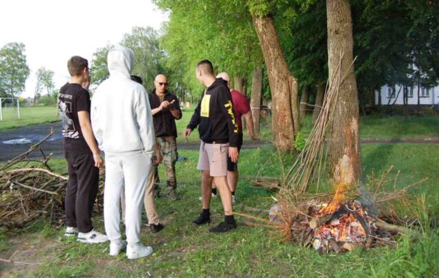 Uczniowie klas mundurowych wraz z opiekunem na terenie boiska szkolnego. Doglądają rozpalonego ogniska. O drzewo oparte są kijki do smażenia kiełbasek.