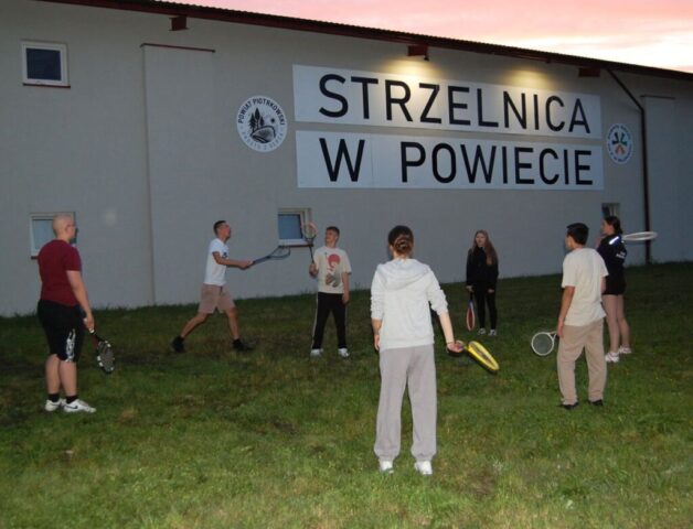 Uczniowie klas mundurowych podczas szkolenia integracyjnego. Stoją na zewnątrz, przy budynku strzelnicy i grają w badmintona.