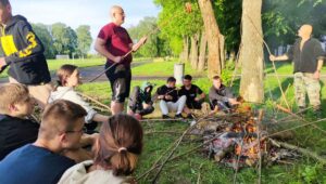 Uczniowie klas mundurowych wraz z opiekunem na zewnątrz, na terenie boiska szkoły. Siedzą na trawie i stoją wokół rozpalonego ogniska. Pieką kiełbaski. W tle bieżnia oraz drzewa. 