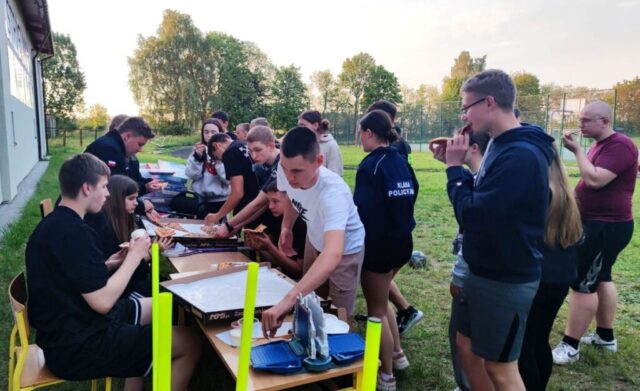 Uczniowie klas mundurowych podczas szkolenia integracyjnego n terenie boiska szkolnego. Siedzą przy ławkach i stoją jedząc kiełbaski. W tle widoczne boisko orlik.