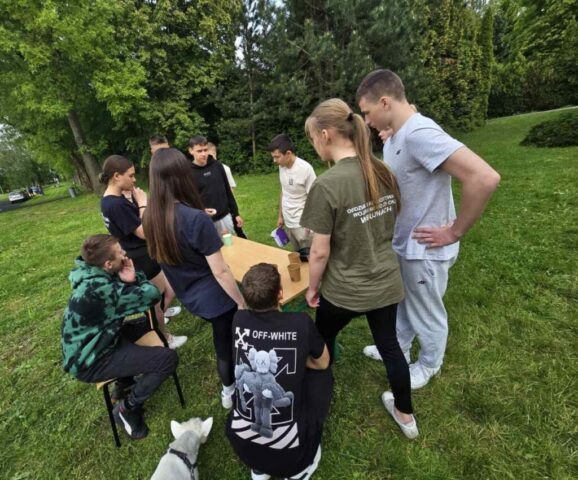 Uczniowie klas mundurowych podczas szkolenia integracyjnego na terenie boiska szkolnego. Siedzą i stoją wokół ławki i rozmawiają. W tle zieleń.