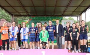 Zwycięskie drużyny sportowe biorące udział w igrzyskach wraz z opiekunami i organizatorem pod wiatą. Pozują do zdjęcia. Zawodnicy mają na szyjach medale. Przed nimi podium. W tle banery reklamowe wydarzenia i organizatorów.