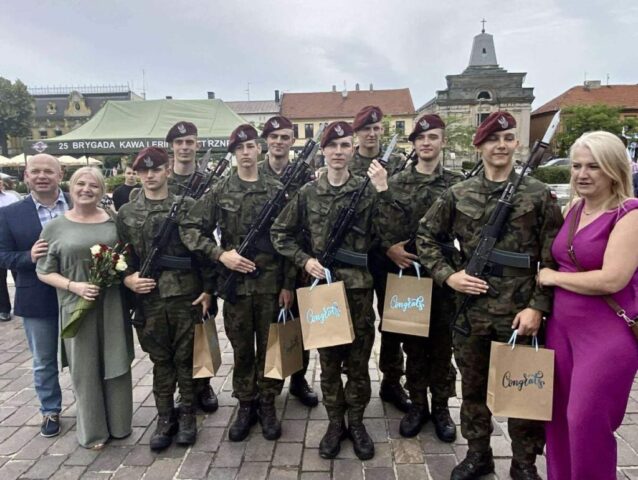 Kadeci OPW, absolwenci ZS CKZ Bujny w umundurowaniu wraz z p. Dyrektor, p. Wychowawczynią i opiekunem klas OPW pozują do zdjęcia w trakcie uroczystości ślubowania po odbyciu I turnusu dobrowolnej zasadniczej służby wojskowej. W tle zabudowania oraz namiot firmowy 25. Brygady Kawalerii Powietrznej.