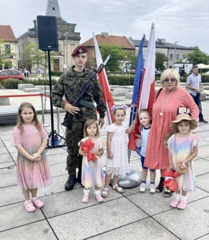 Kadet klasy OPW, absolwent szkoły w umundurowaniu pozuje do zdjęcia w towarzystwie dzieci oraz kobiety. Stoi przy stojaku z Flagami Polski oraz flagą koloru granatowego. W tle zabudowania.