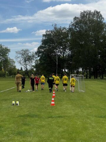 Uczestnicy wieloboju sportowego oraz przedstawiciele służb mundurowych na boisku szkoły. widać fragment toru przeszkód, czerwono-białe pachołki i bramkę.
