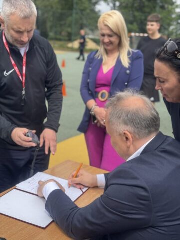 Stolik sędziowski. Pan Wicemarszałek dokonuje zapisu wyniku, który odczytuje ze stopera trzymanego przez nauczyciela WF. Widoczna także Pani Dyrektor Szkoły oraz Pani Prezes Luks Gryf. W tle boisko orlik.
